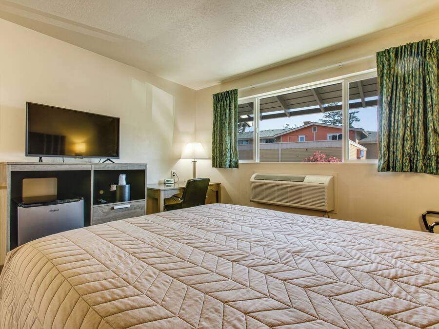 A hotel room with a large bed, a desk with a computer, a television, a mini-fridge, and a window with curtains.