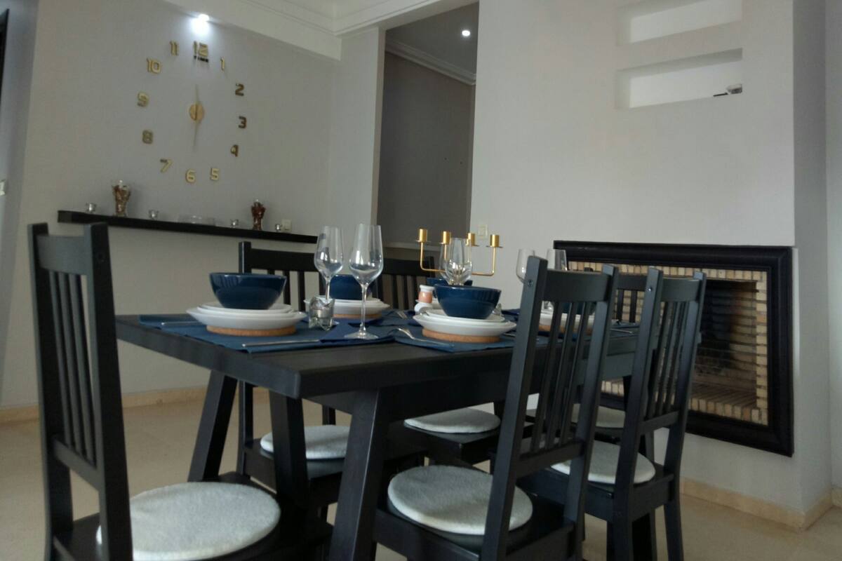 A dining area with a table set for four, chairs, a chandelier, and a wall clock.