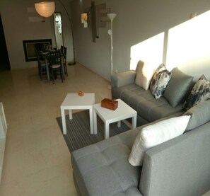 A modern living room with a grey sofa, two white coffee tables, and a dining area with a table and chairs.