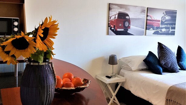 A hotel room with a bed, a bedside table with a lamp, a vase with sunflowers and oranges, and two wall-mounted car posters. - 12 Simonsplein (Stellenbosch)