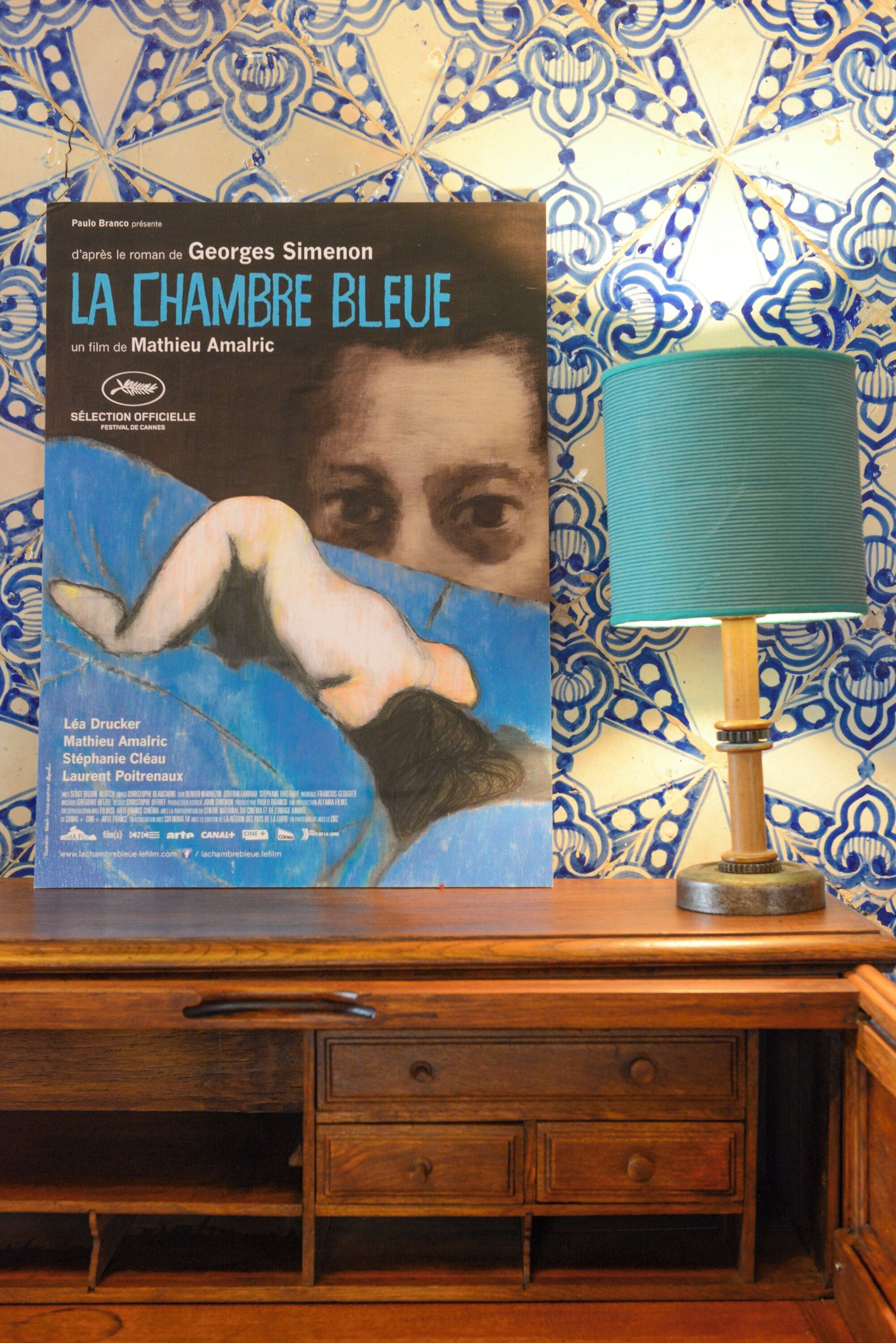 Un cartel de película titulado "LA CHAMBRE BLEUE" de Georges Simenon, sobre un escritorio de madera frente a una pared con diseño.