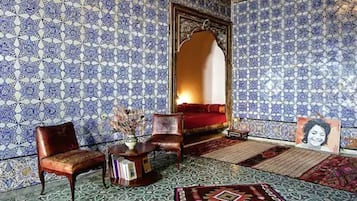 Una habitación con pared de azulejos blancos y azules con diseño, un sofá rojo, una silla de madera, una mesita con un florero y un espejo grande.