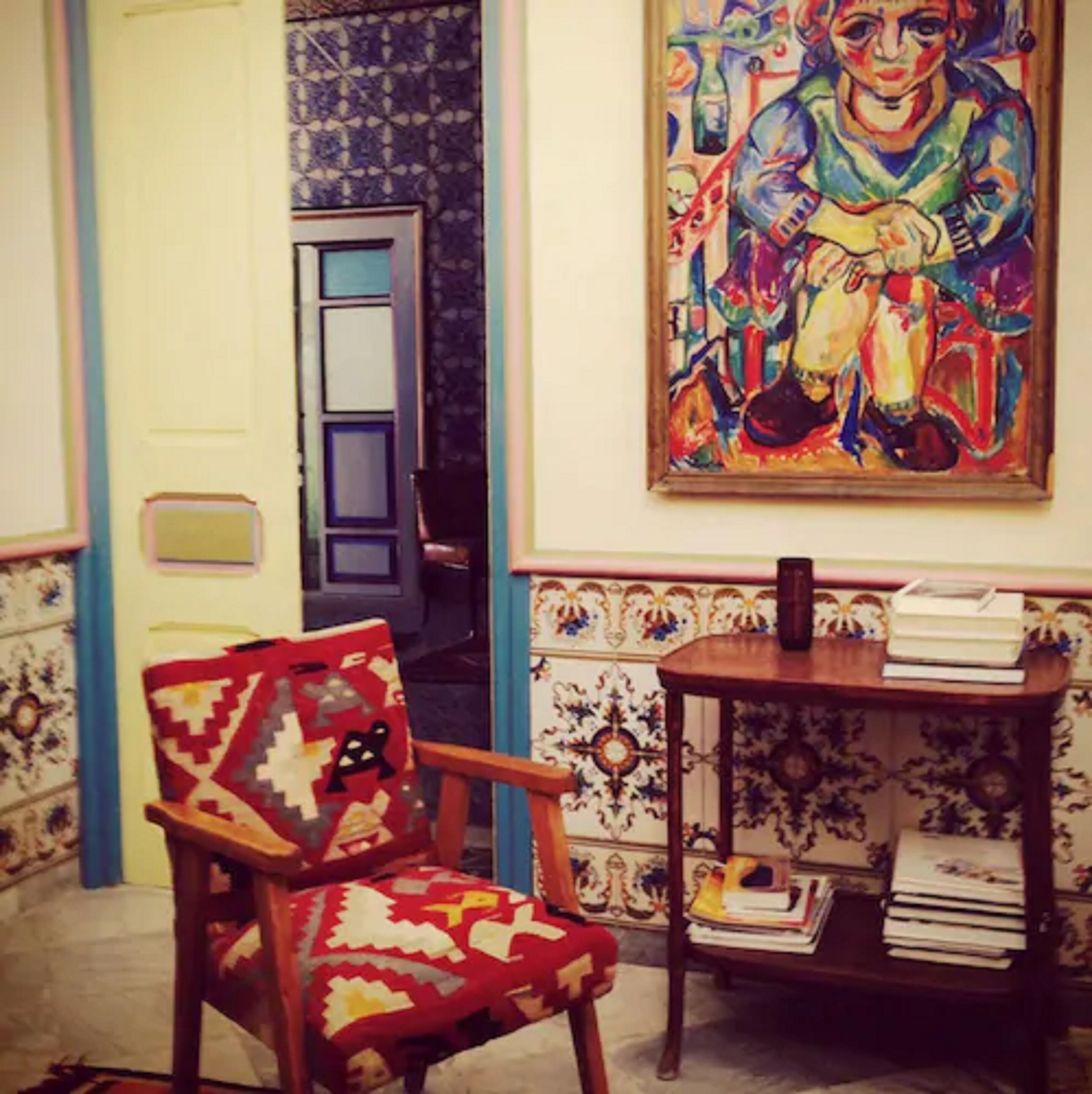 Una habitación con pared estampada, una silla de madera, una mesa con libros y un cuadro colorido.