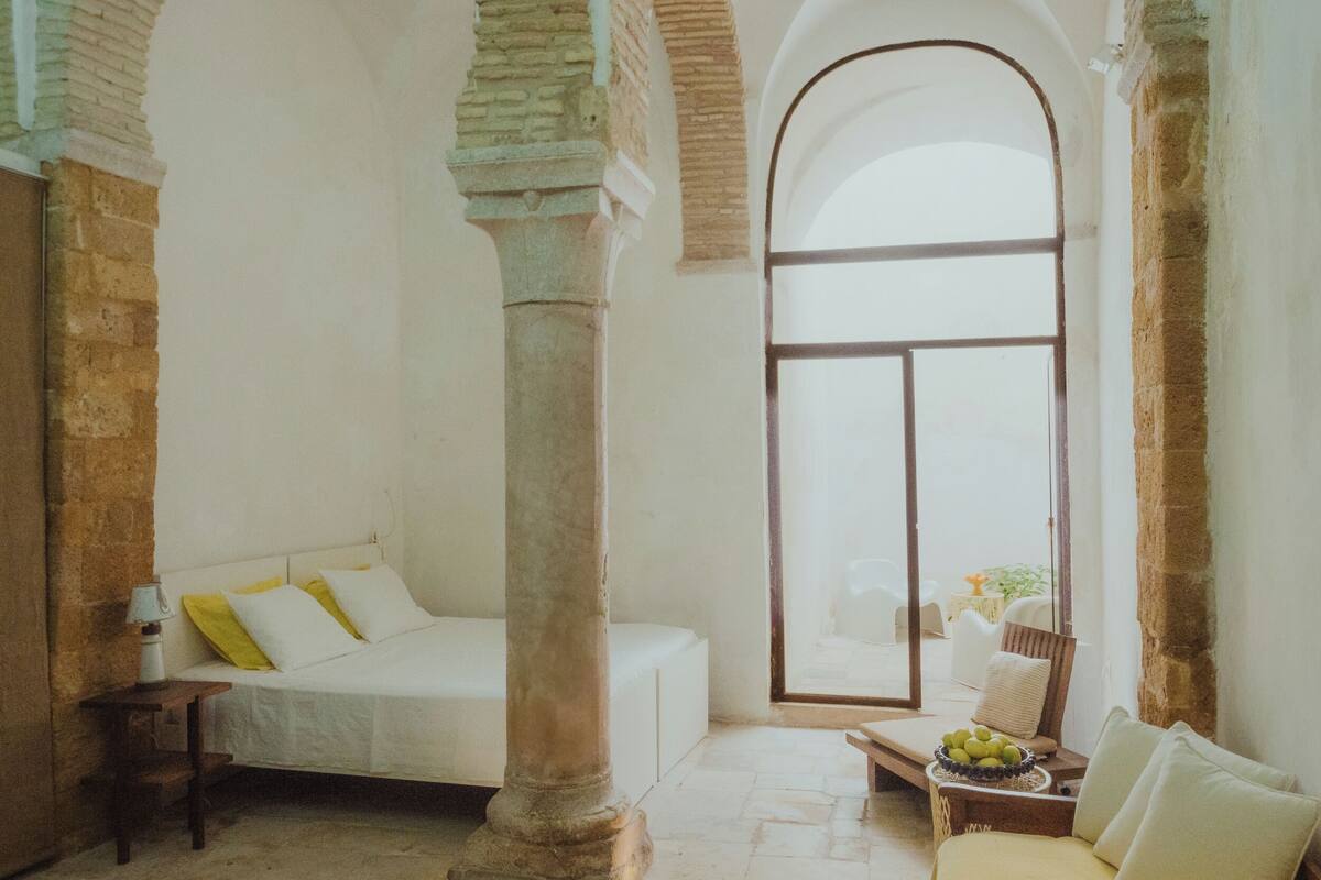 A room with a bed, a table with fruit, and a view through a large window.