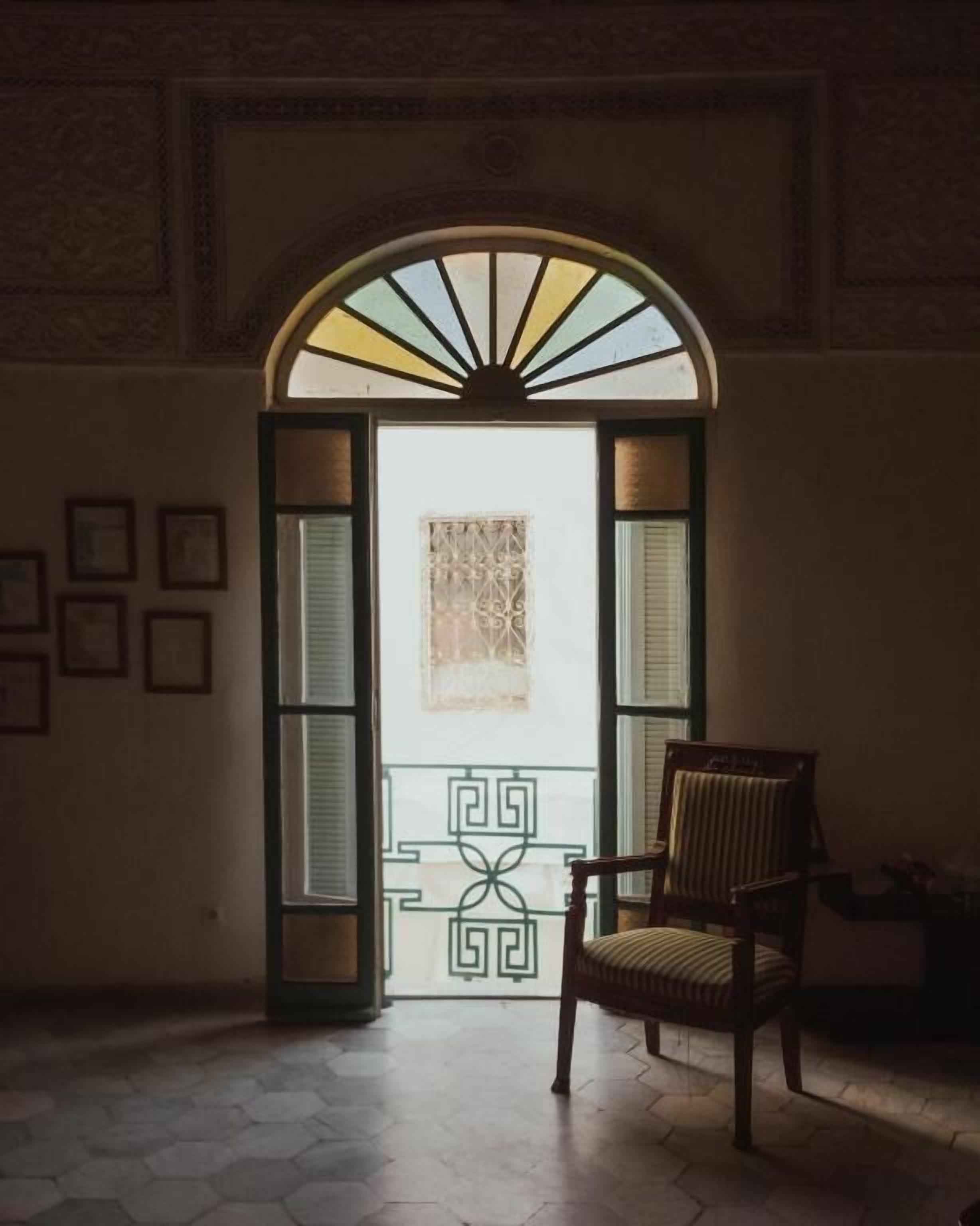 Una habitación interior con un amplio arco que da a un balcón, una silla y un ventanal con diseño.