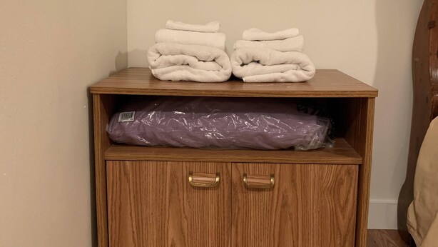 Un meuble en bois avec deux tiroirs et une étagère, contenant des serviettes blanches pliées et un sac en plastique violet.