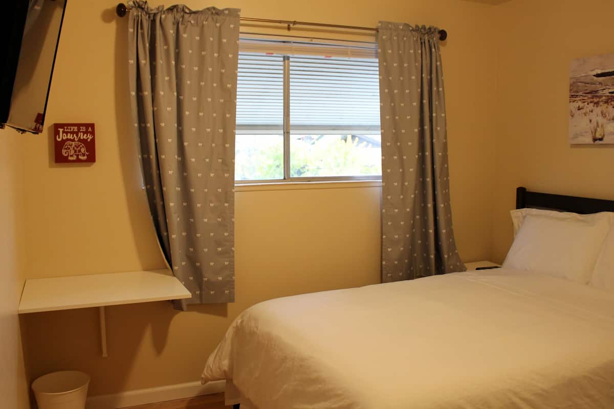 A bedroom with a bed, a window with curtains, a bedside table, and a wall-mounted shelf.