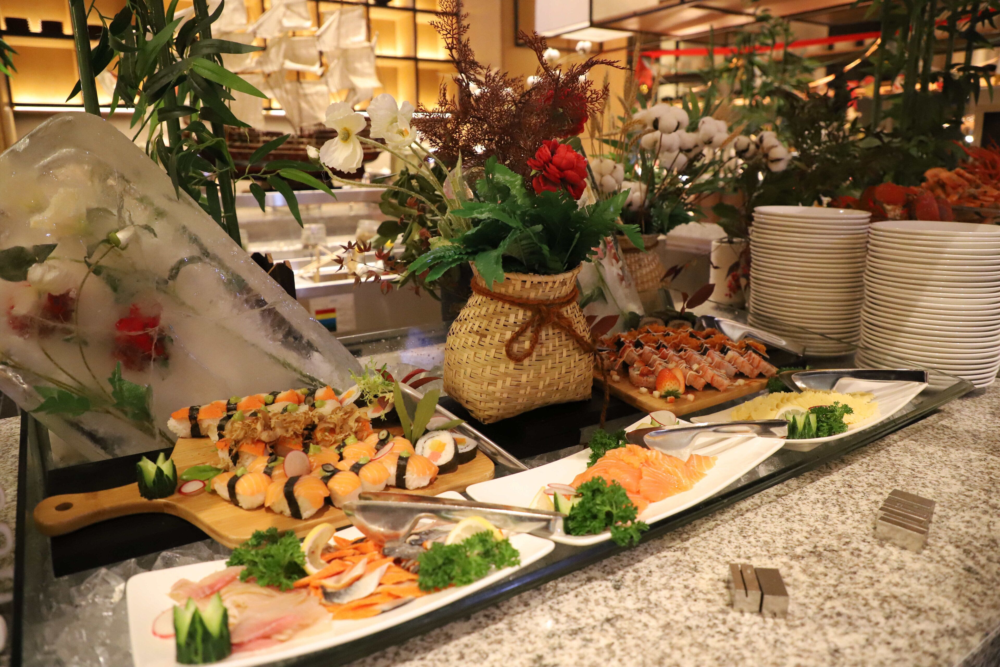 A buffet with various dishes, including sushi and seafood, arranged on a counter with a decorative plant in the background.
