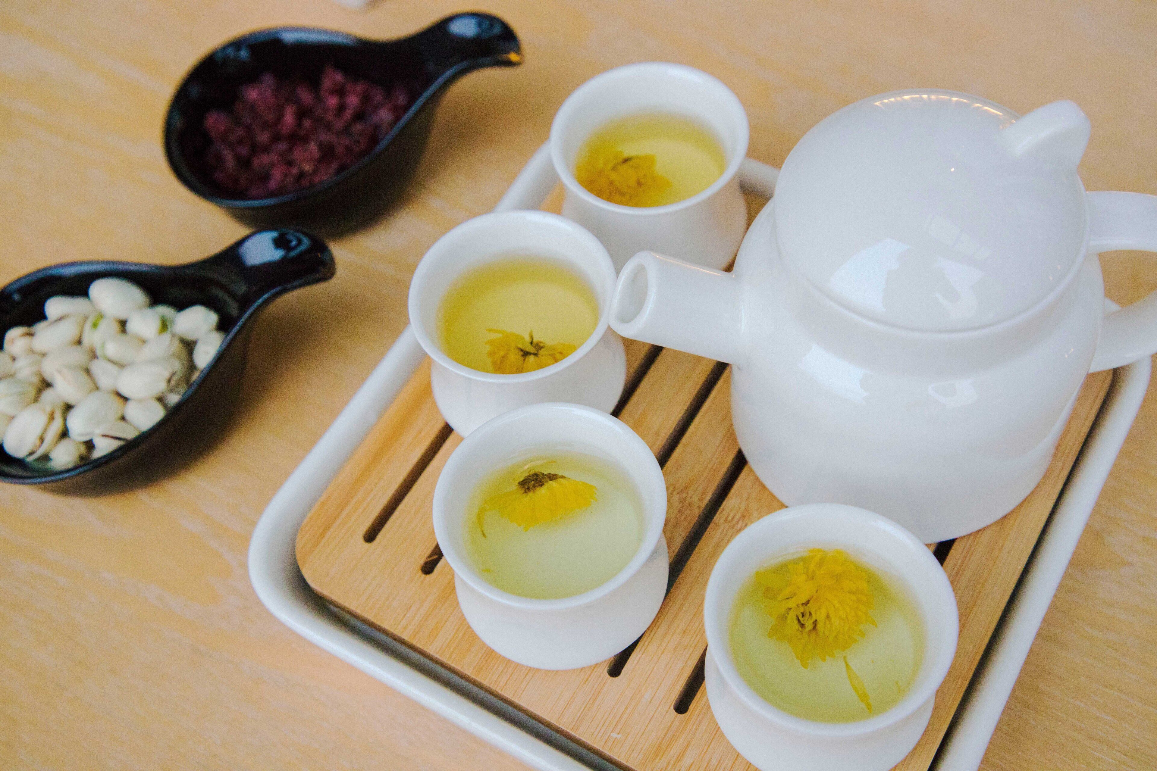 A tray with a white teapot and four cups of tea, each with a yellow flower, and a small black dish with white beans.
