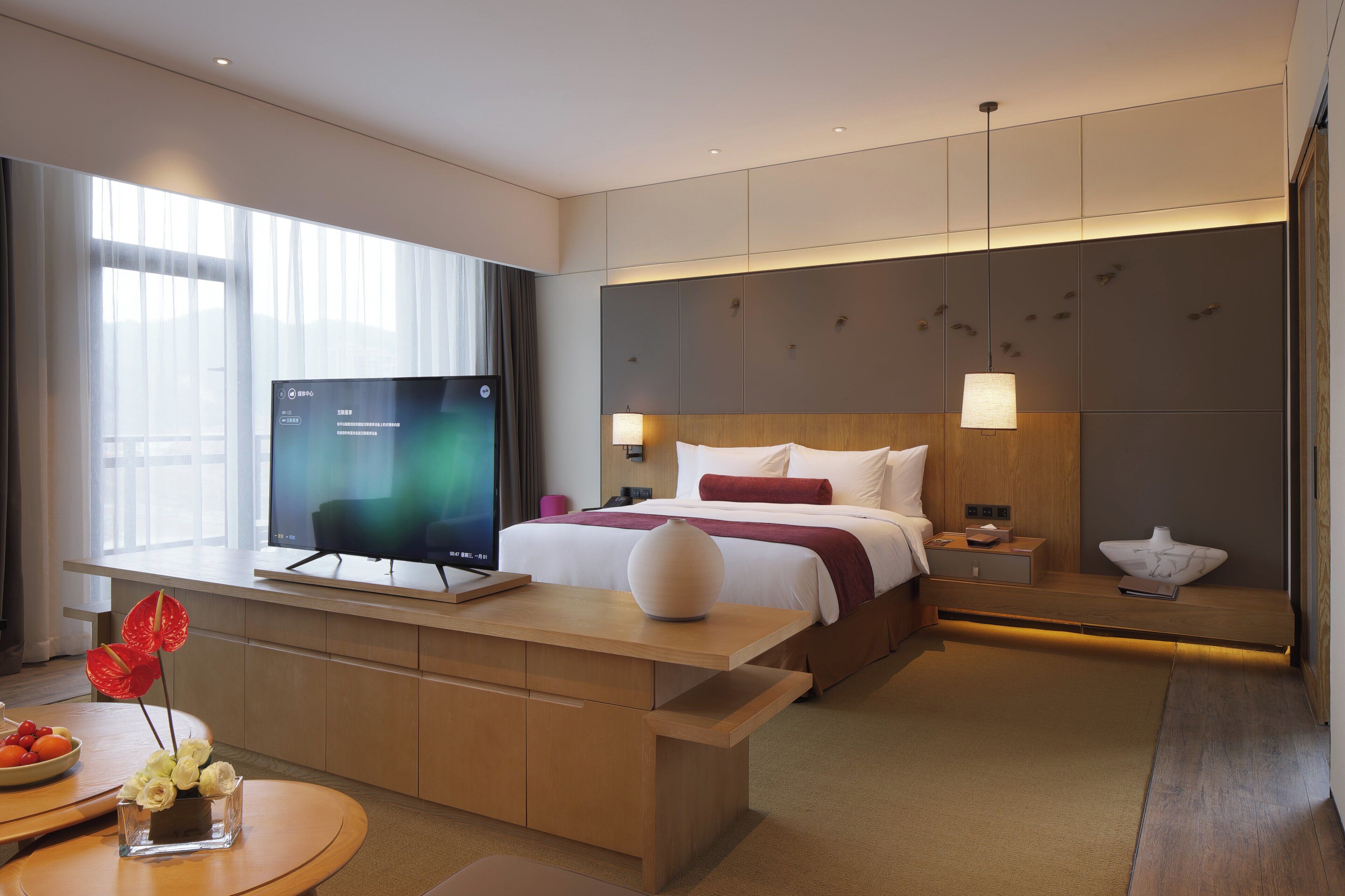 Een hotelkamer met een bed, nachtkastje, televisie, en een bureau met stoel en een vaas bloemen.