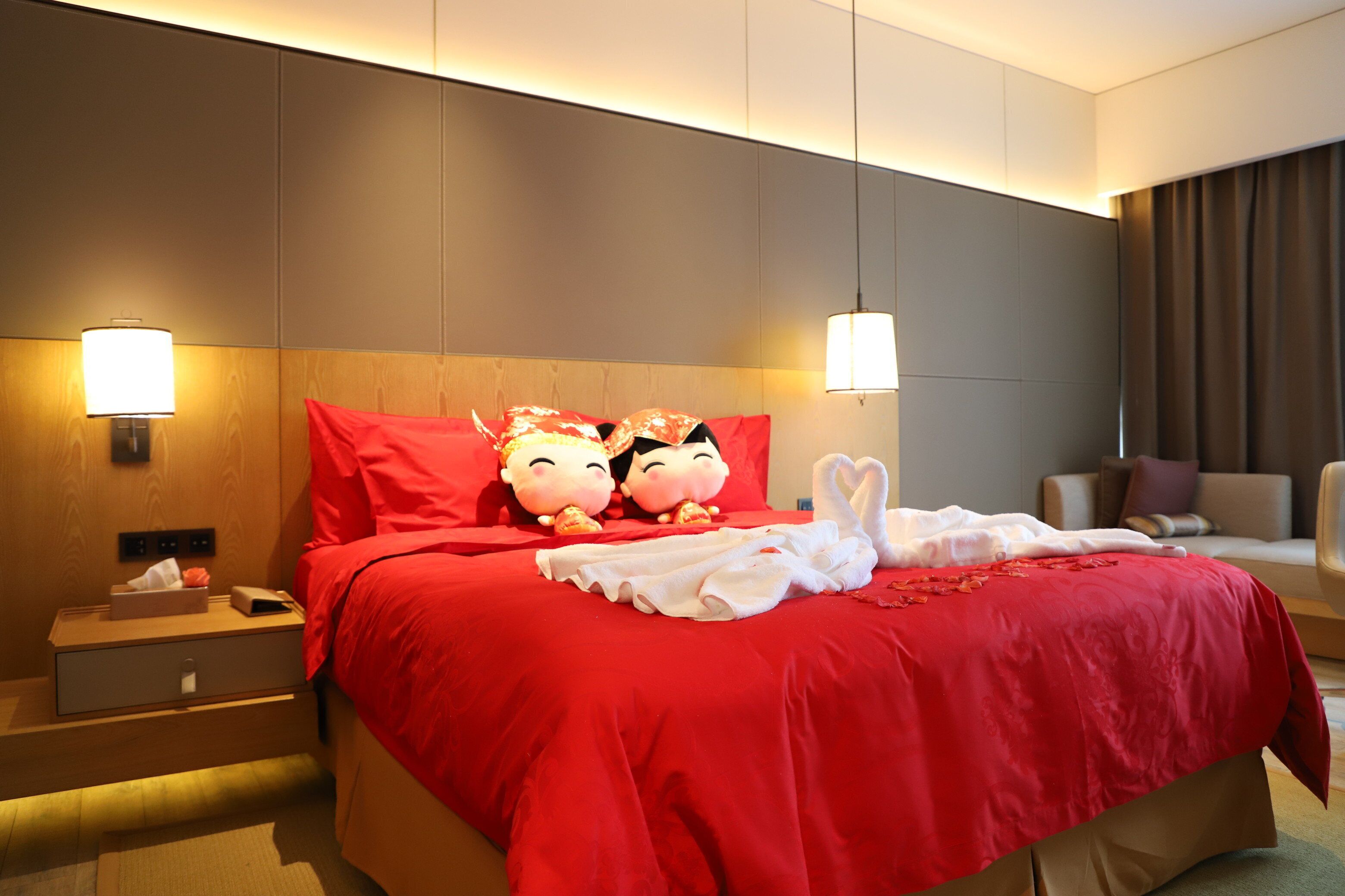 A hotel room with a large bed, red bedding, two wall-mounted lamps, a nightstand with a lamp, a small table with a tissue box, and a sofa.