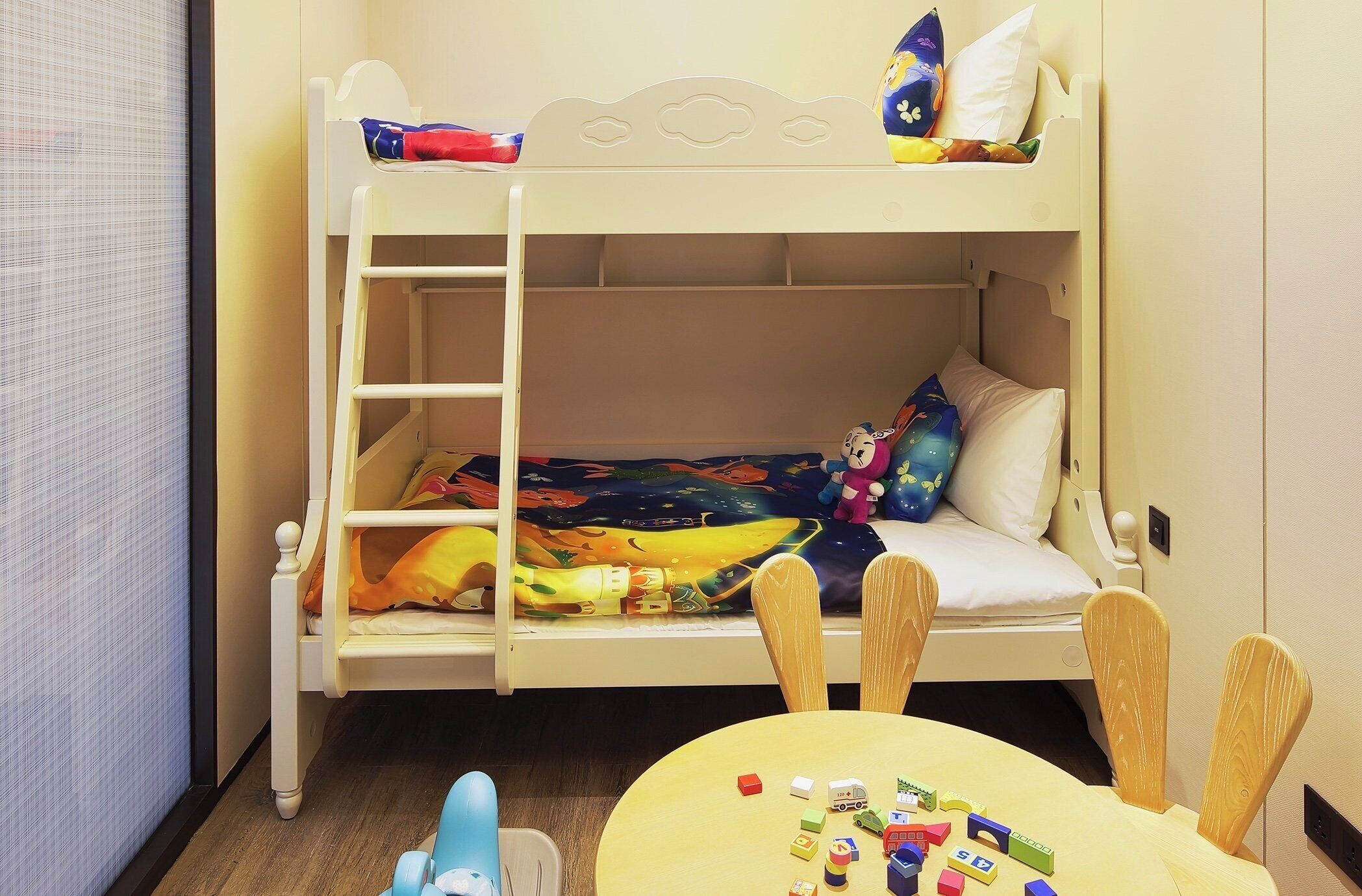 Una cama litera con escalera, una mesa con juguetes y una ventana con persianas.