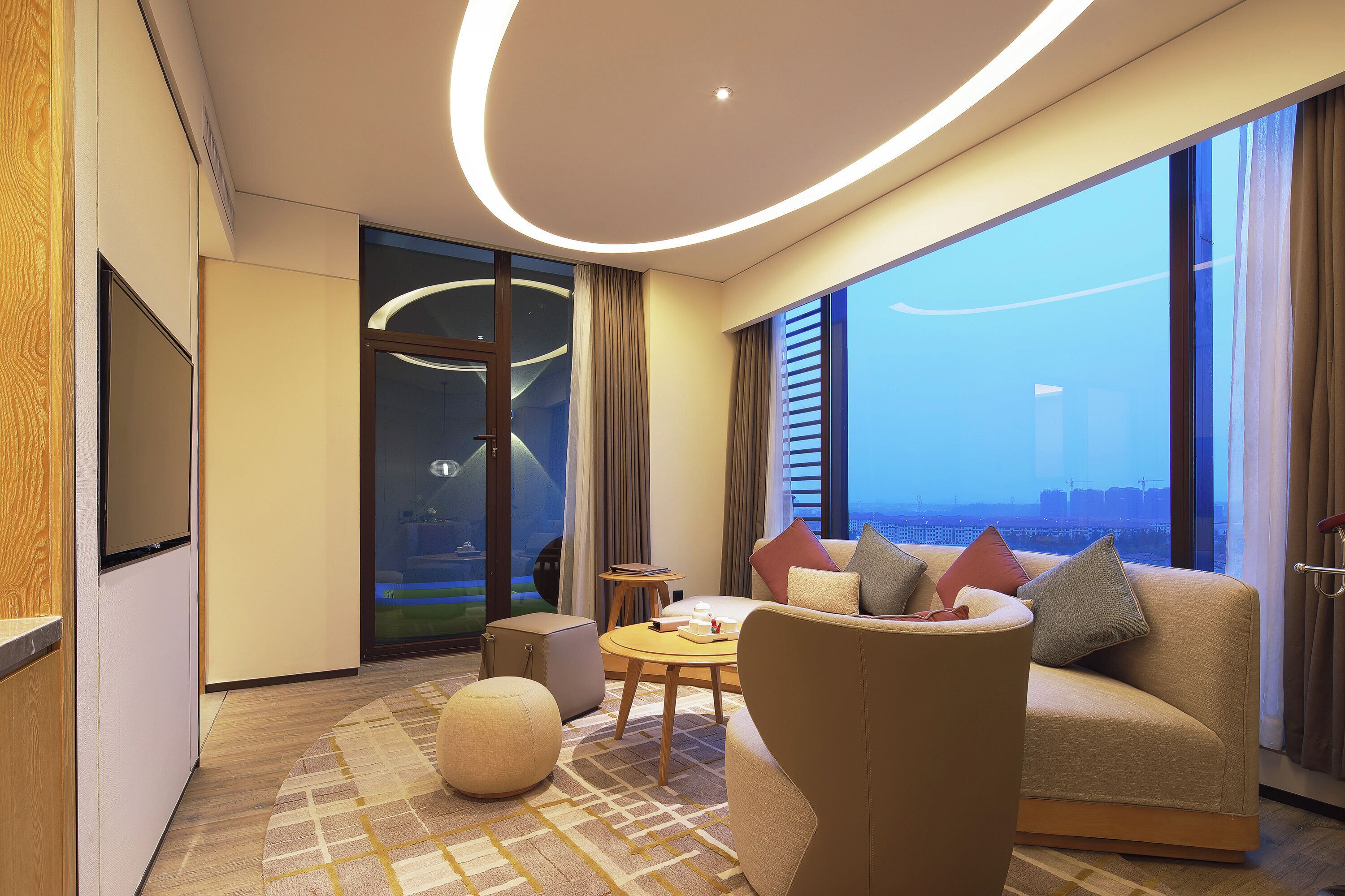 A modern hotel room with a sofa, a small round table, a TV, and a large window offering a city view.
