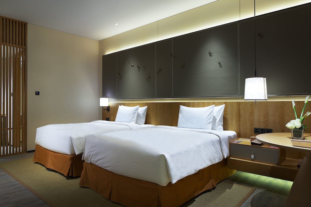 Habitación de hotel con dos camas, cabecero de madera, mesita de noche con lámpara y un pequeño escritorio con un florero.