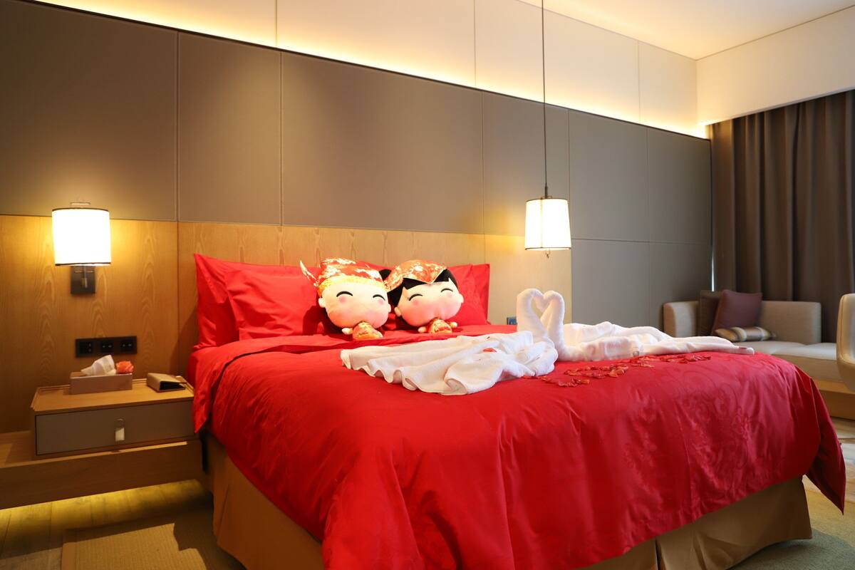 Habitación de hotel con una cama grande, dos almohadones decorativos, una mesita de noche con lámpara y otro mueble auxiliar.