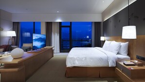 Habitación de hotel moderna con una cama grande, un escritorio, un televisor y vistas de la ciudad de noche.
