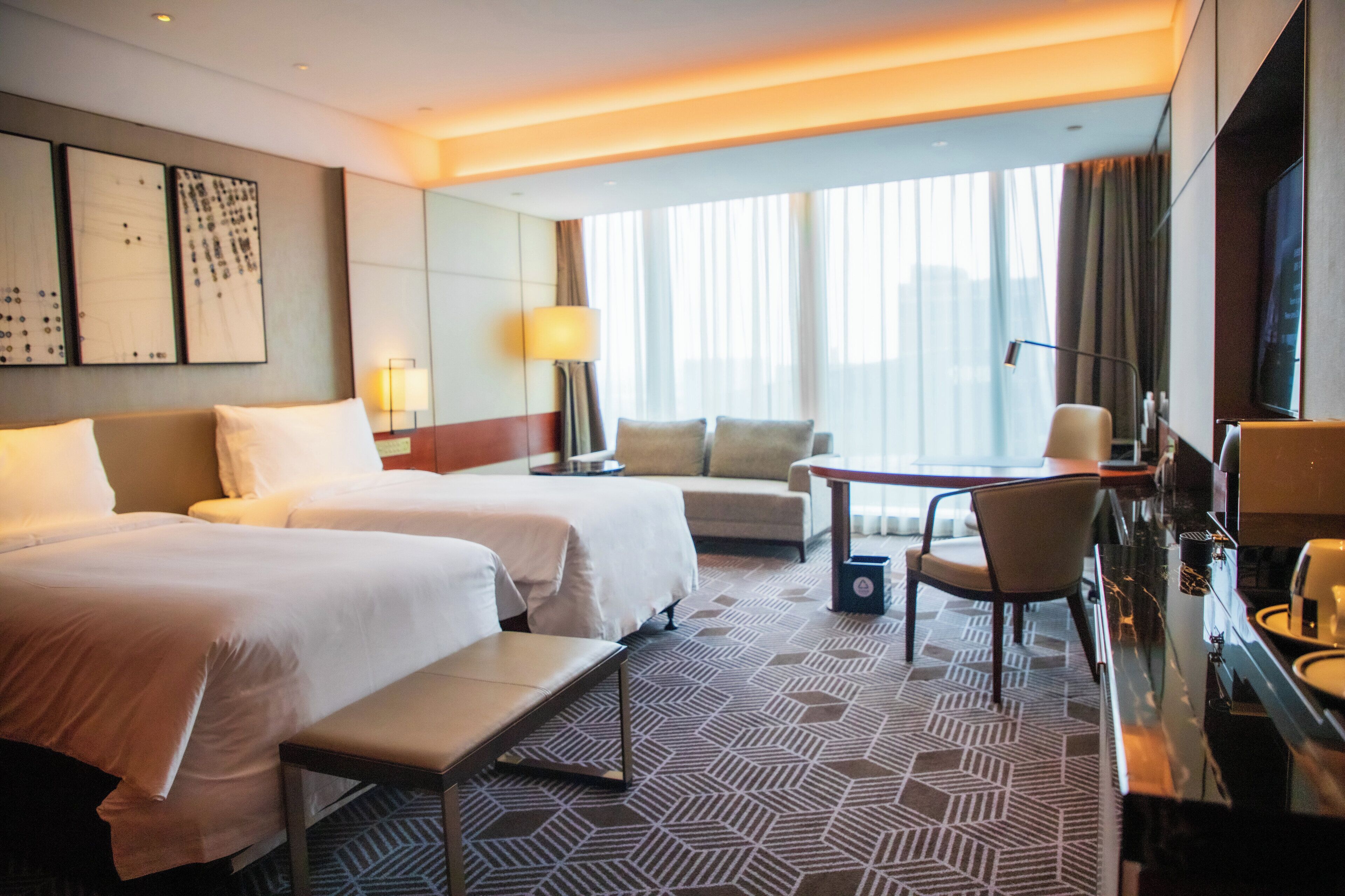 A hotel room with two beds, a sofa, a desk, a chair, and a large window with curtains.