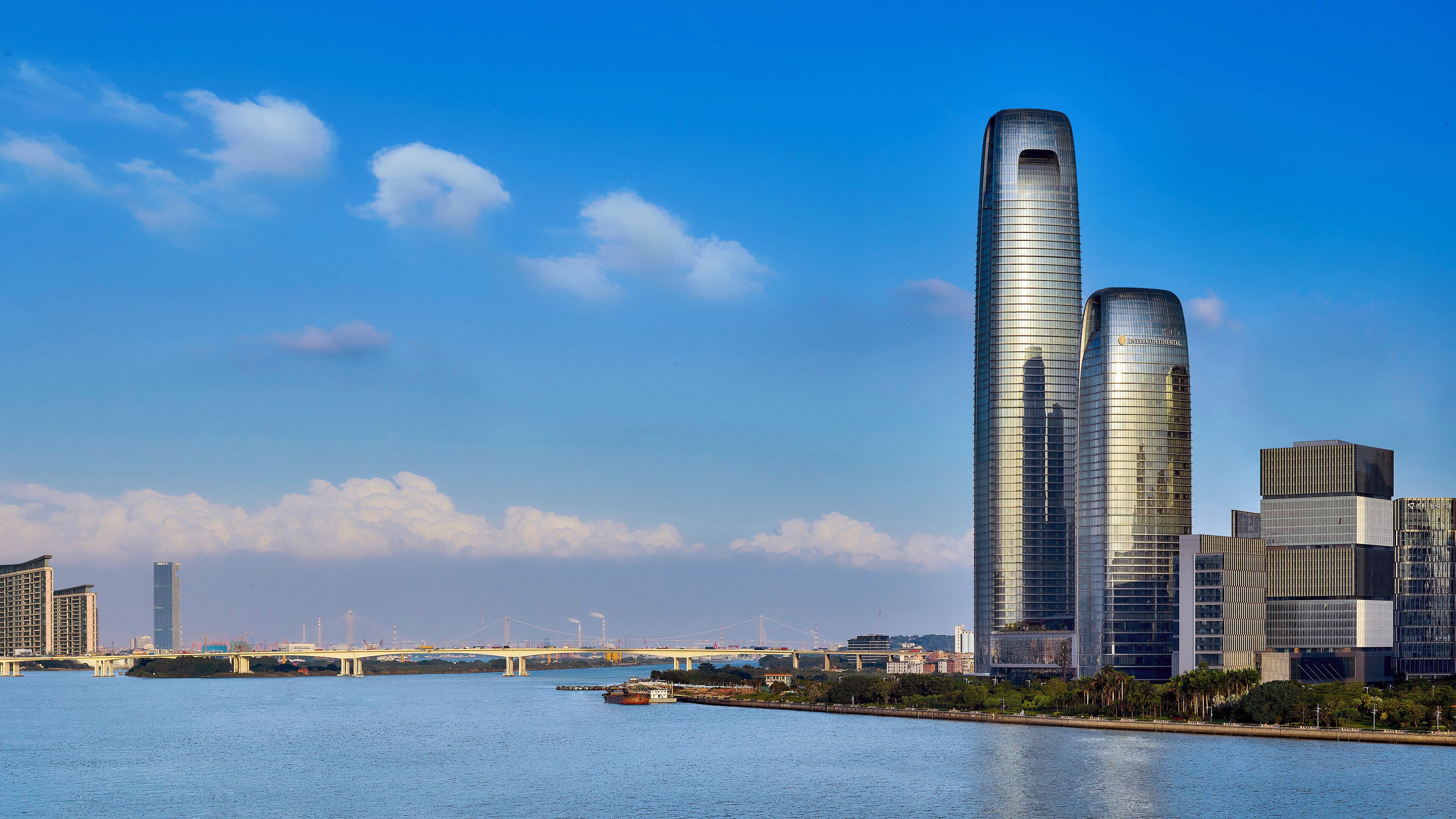 A modern high-rise building with a unique curved design, situated by a river with a bridge in the background.