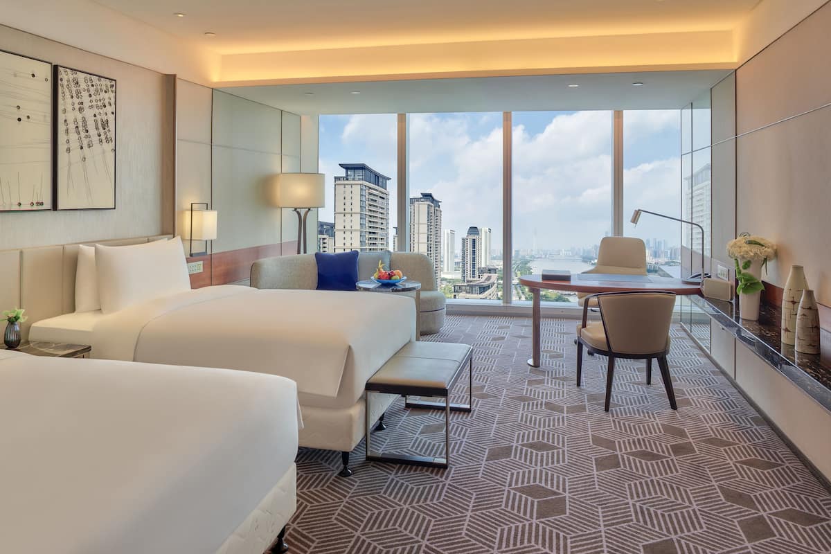 A hotel room with two beds, a desk, a chair, and a large window with a city view.