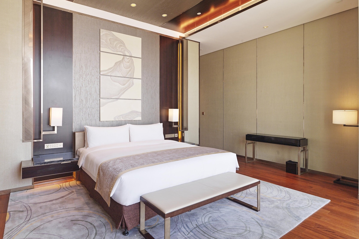 Una habitación de hotel moderna con una cama grande, mesitas de noche, un banco y una obra de arte abstracta de gran tamaño en la pared.