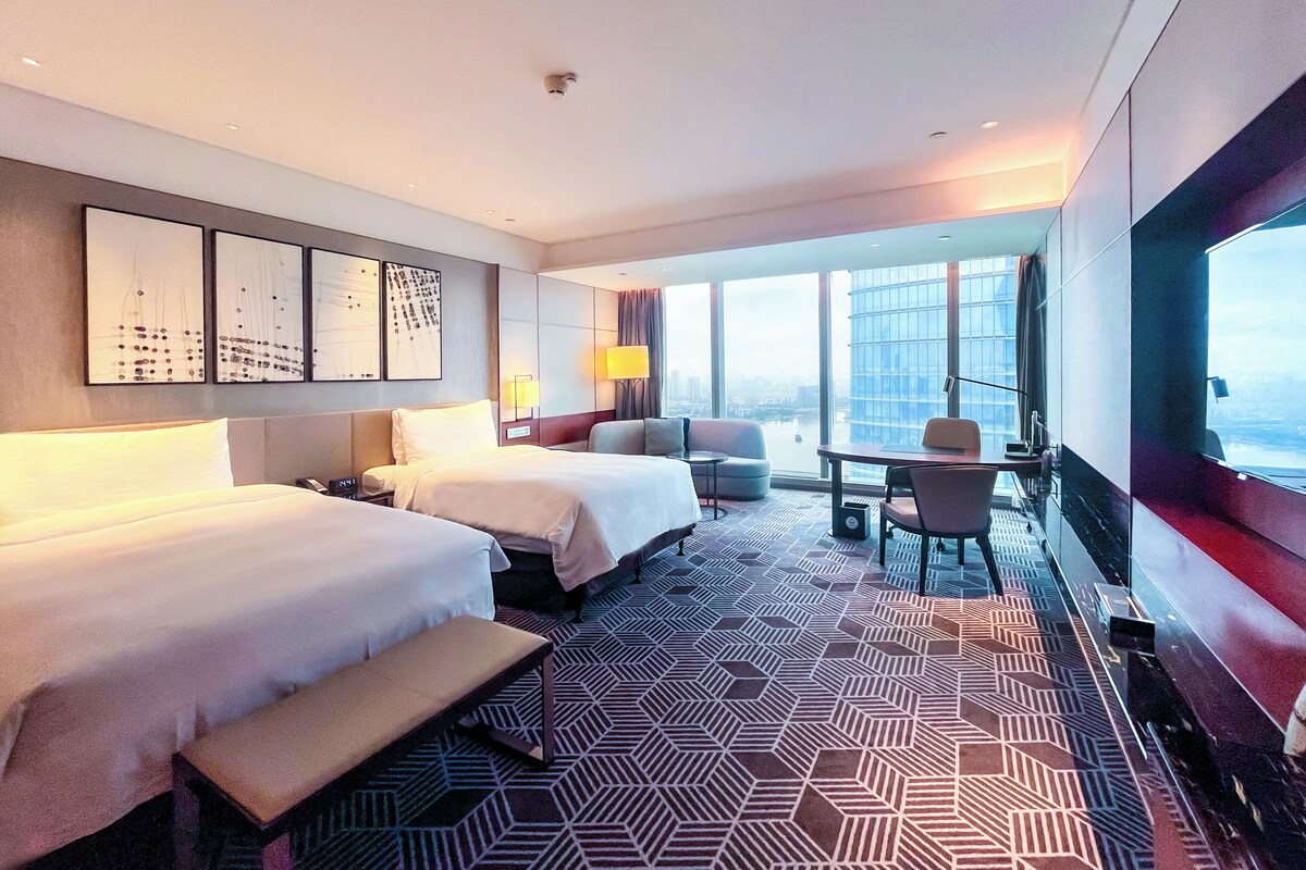 A hotel room with two beds, a desk, a chair, and a large window offering a city view.