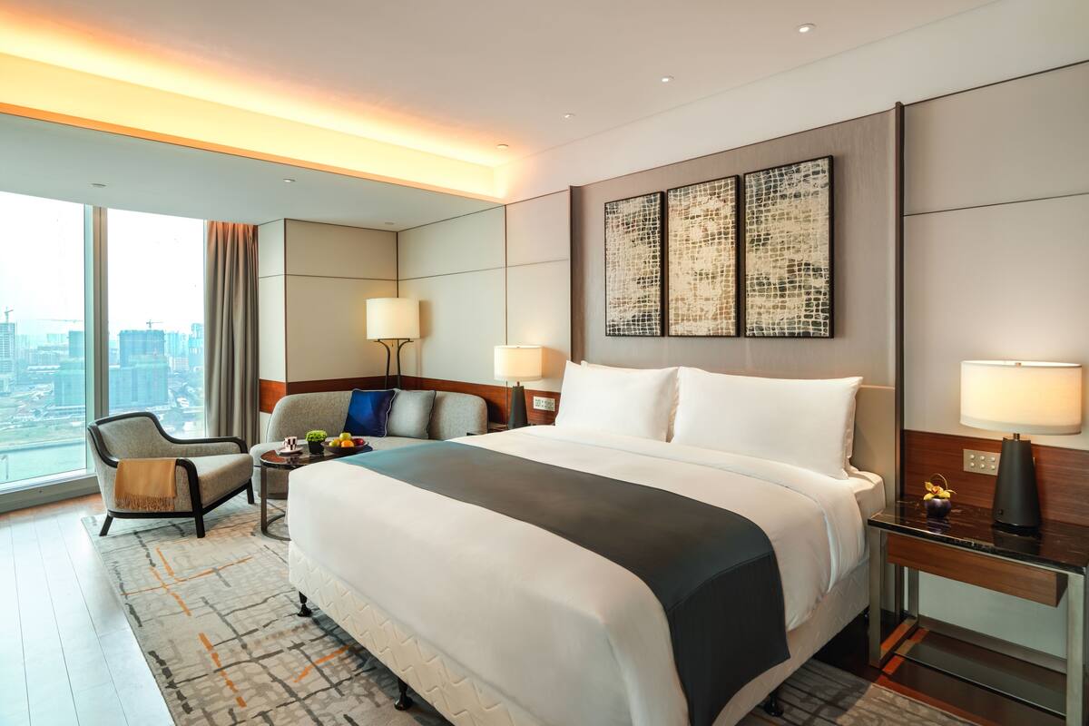 A modern hotel room with a large bed, a seating area with a chair and table, a desk with a lamp, and a city view through large windows.