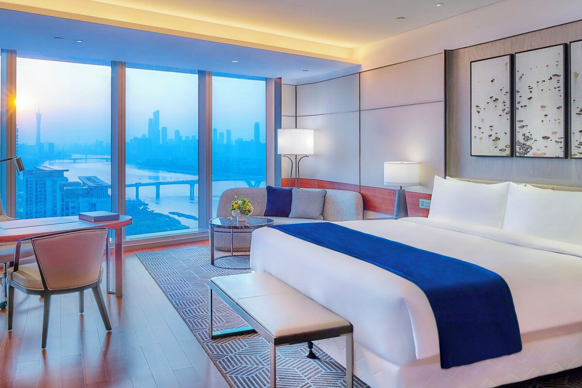 A hotel room with a large bed, a desk, a chair, a small table with a vase of flowers, and a view of a cityscape through large windows.