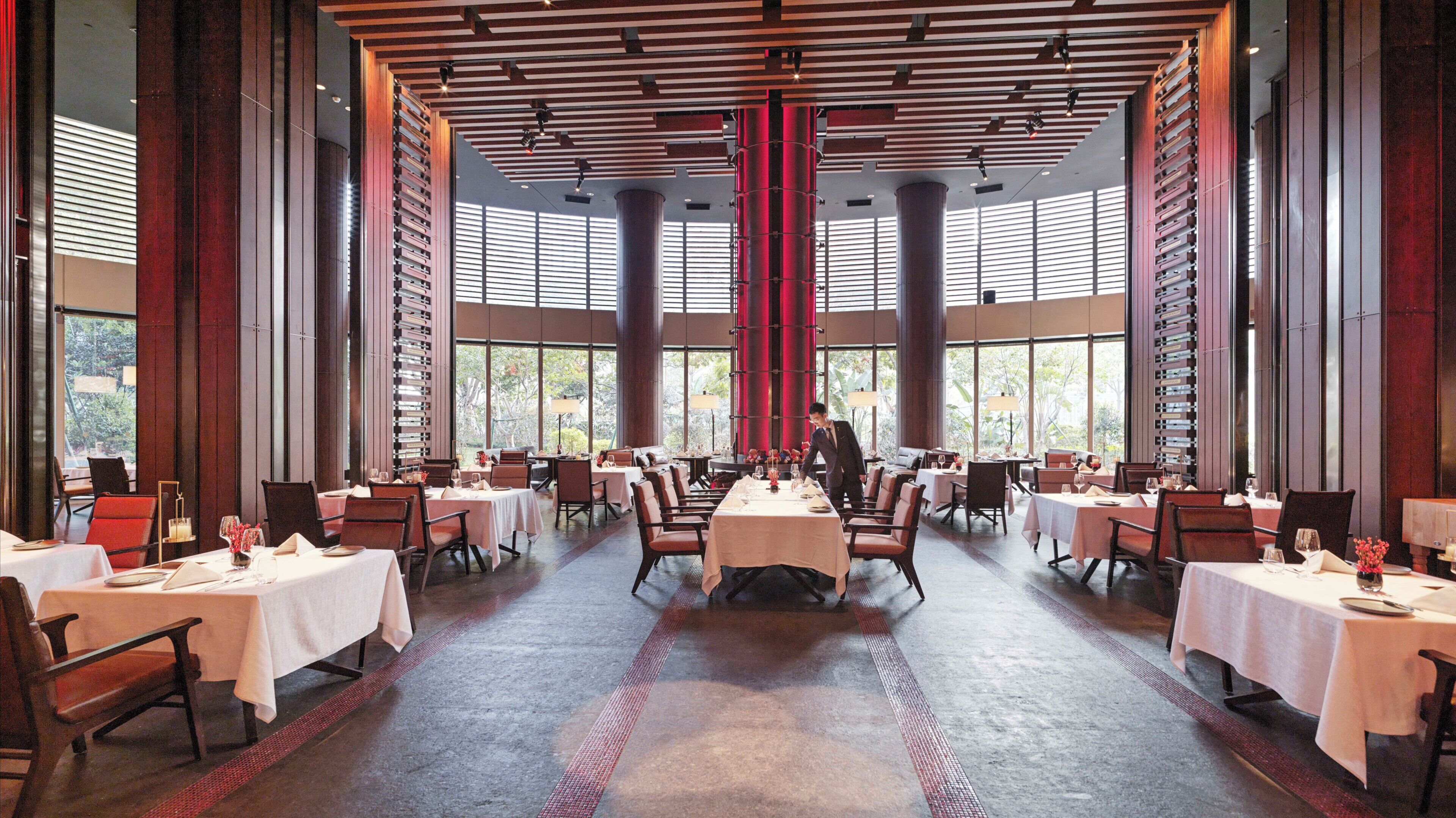 A spacious dining area with multiple tables and chairs, large windows, and a high ceiling with exposed beams.