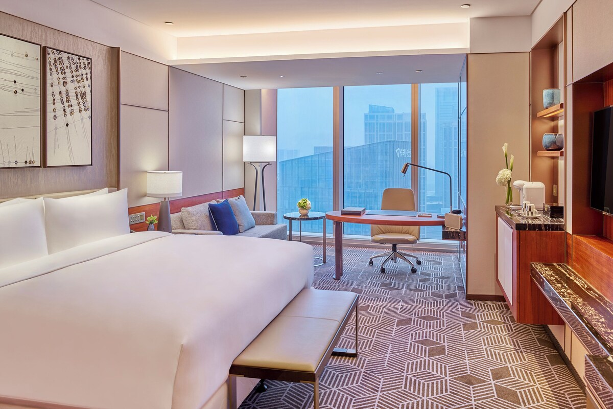 A hotel room with a large bed, a desk with a chair, a TV, and a view of the cityscape.