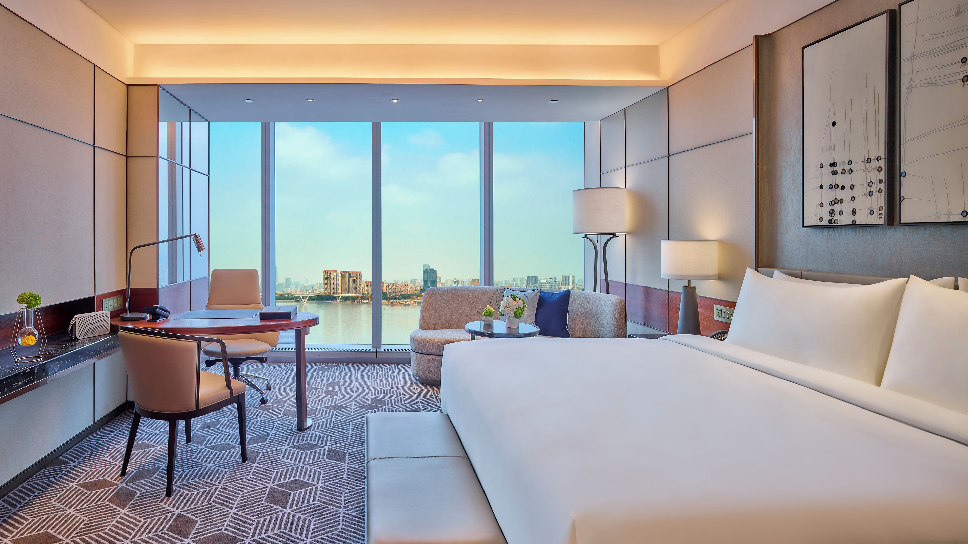 Habitación de hotel con una cama grande, un escritorio con silla, una mesita de noche con lámpara y vista a un paisaje urbano a través de amplias ventanas.