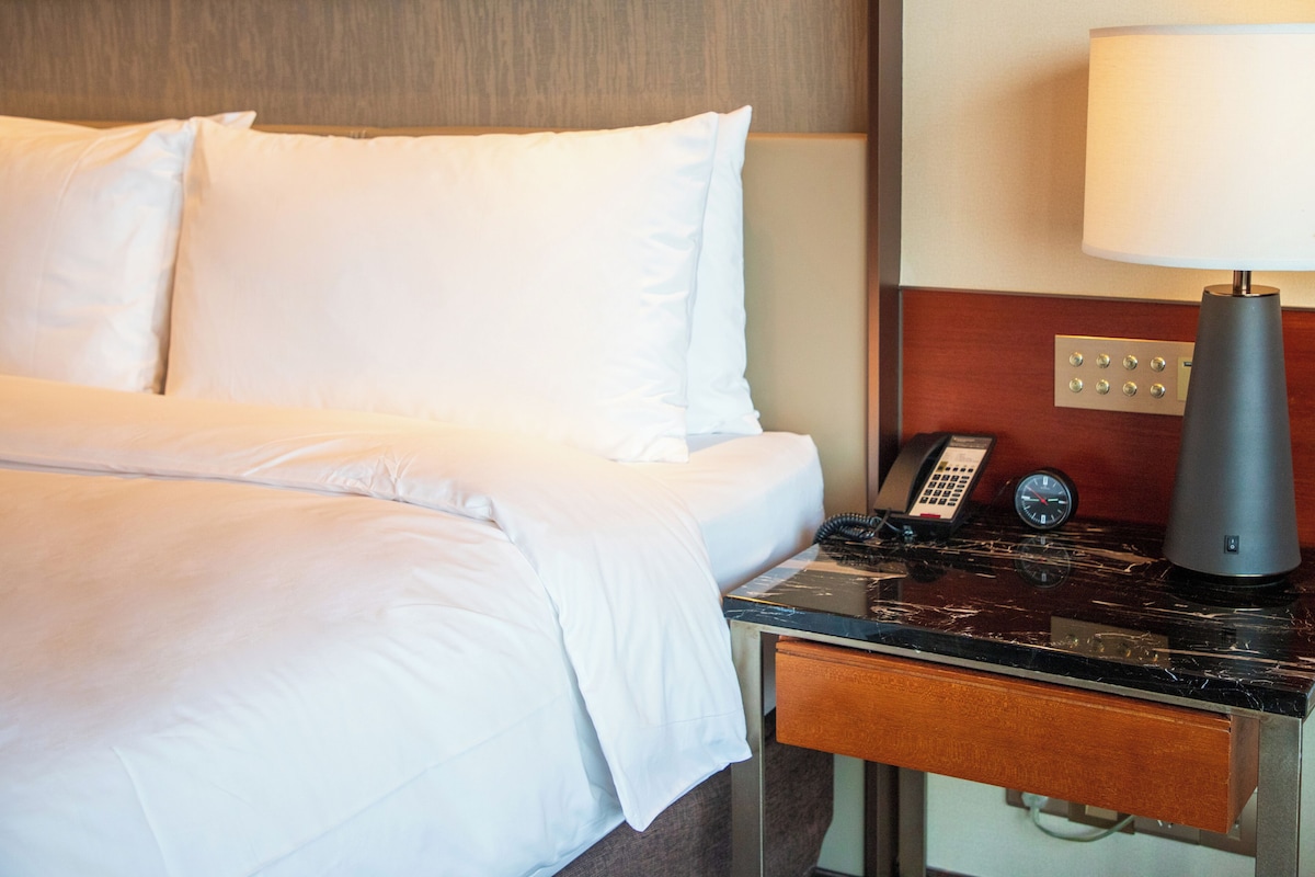A hotel room with a bed, a bedside table with a lamp, a phone, and a clock.