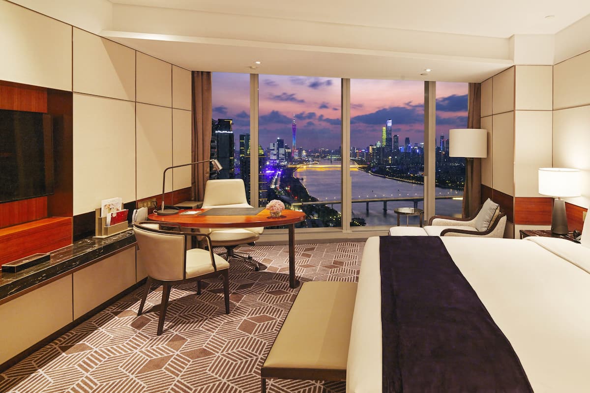 A hotel room with a large bed, a desk with a chair, a small table, and a view of a cityscape at dusk.