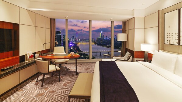 A hotel room with a large bed, a desk with a chair, a small table, and a view of a cityscape at dusk.