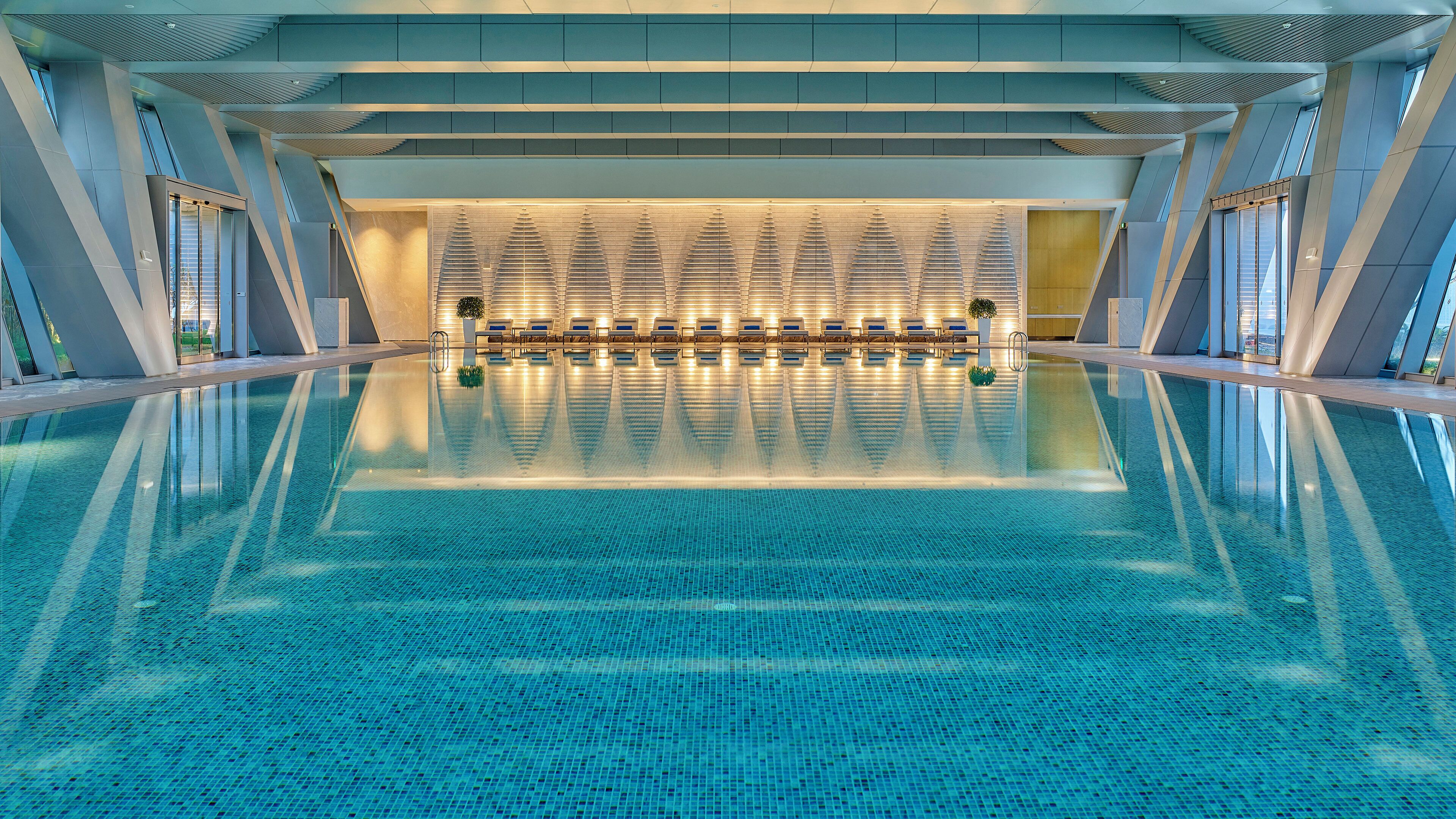 Una amplia piscina cubierta con diseño moderno, que cuenta con una zona central de descanso con tumbonas y un techo con estampado.
