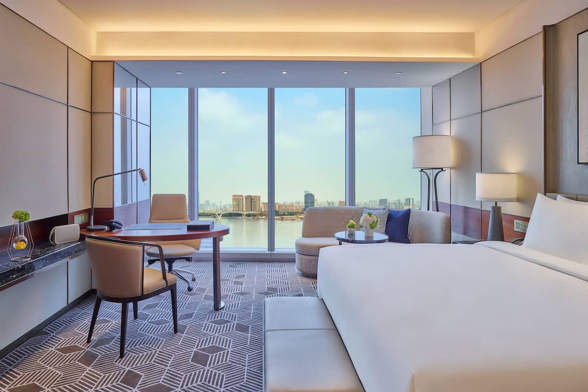A modern hotel room with a large bed, a desk with a chair, a sofa, and a view of a cityscape through large windows.