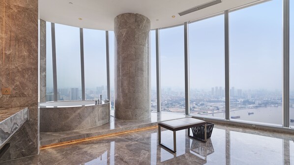 A high-rise hotel room with a large bathtub, a marble floor, and a view of a cityscape through large windows.