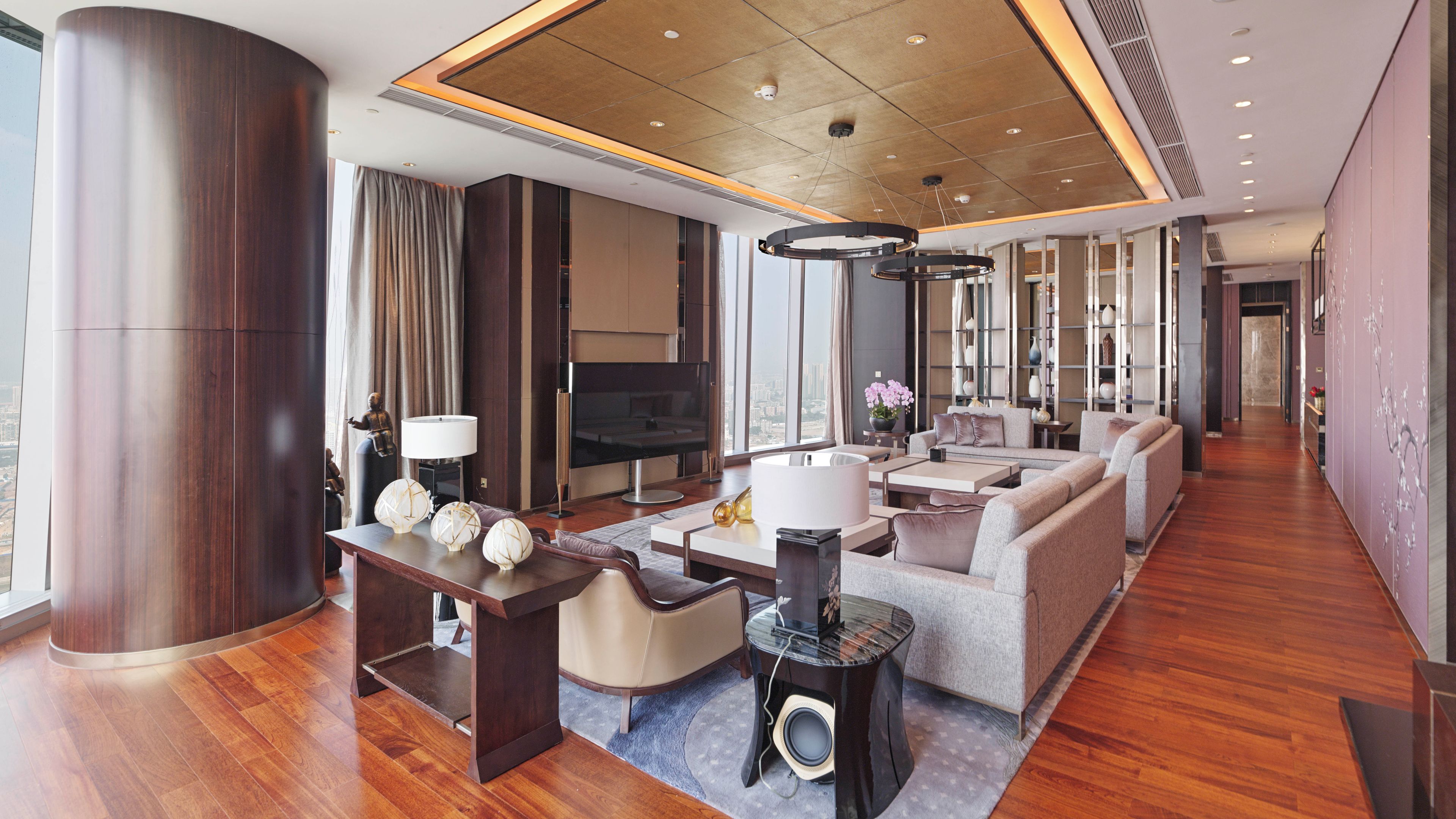 A spacious living area with a large sofa set, a coffee table, and a television. There is a large pillar and a modern ceiling design.