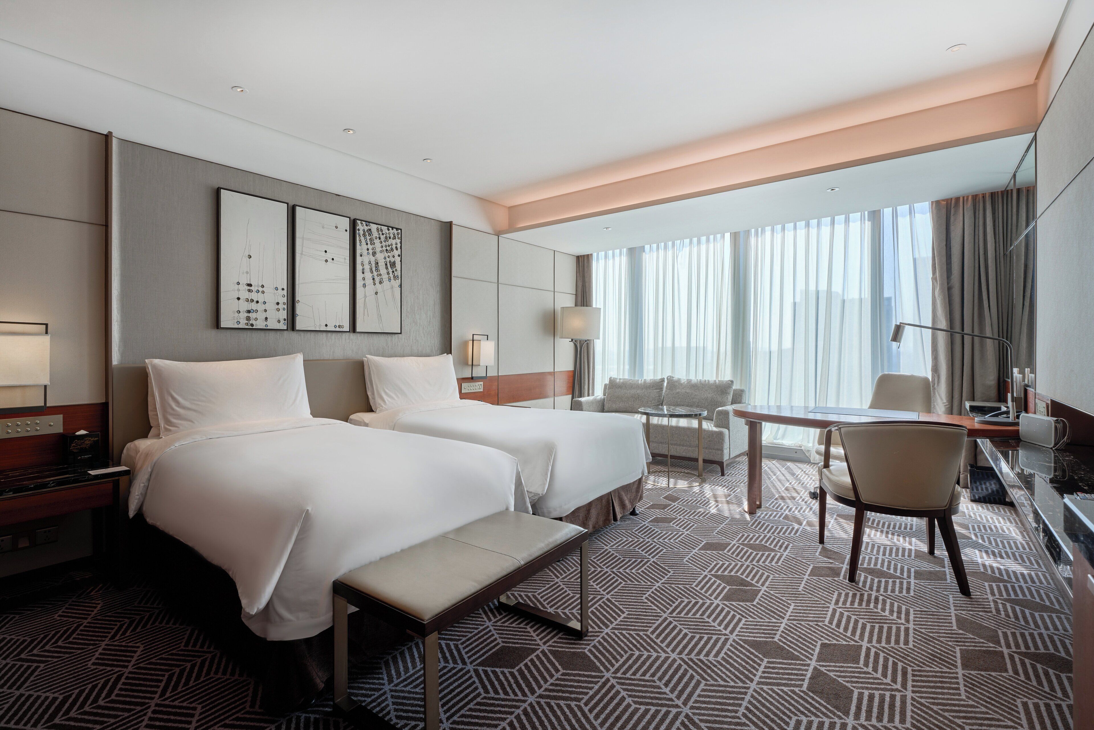A hotel room with two beds, a desk, a chair, and a large window with curtains.