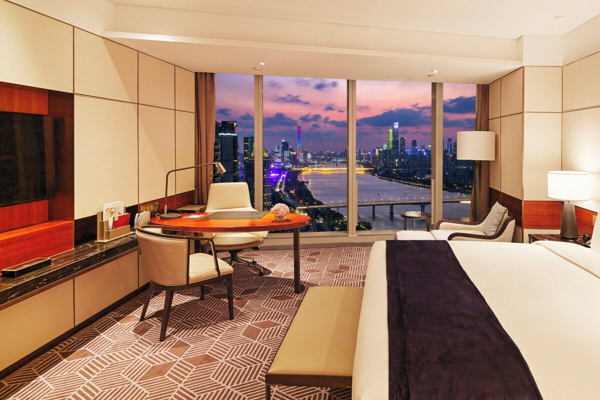 A hotel room with a large bed, a desk with a chair, a TV, and a view of a cityscape at dusk.