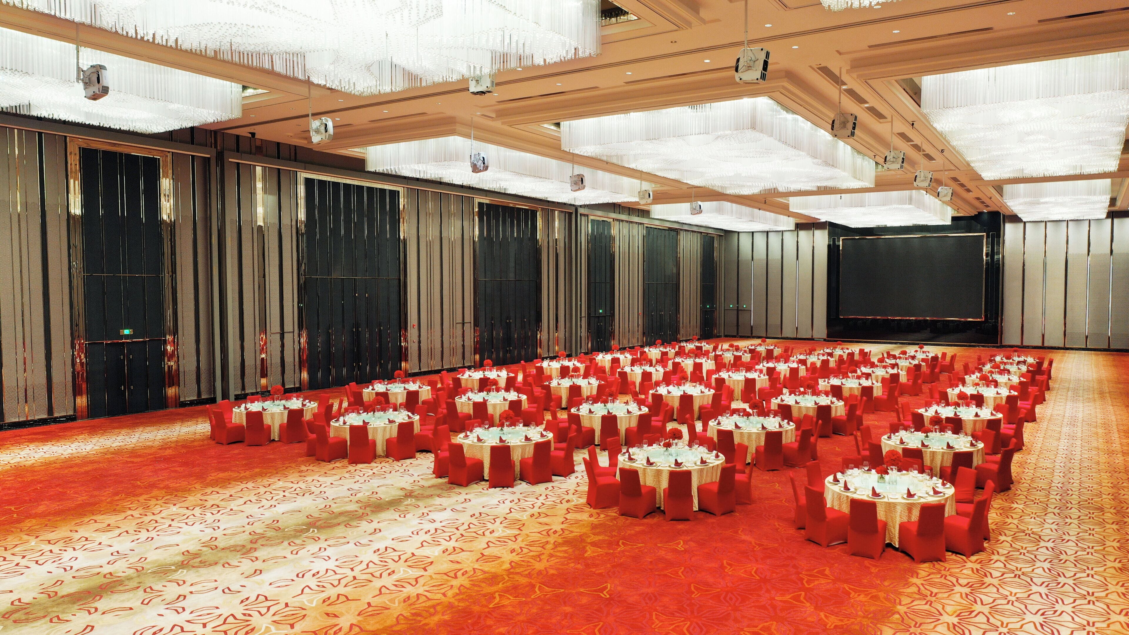 Une grande salle avec des rangées de tables rondes et des chaises rouges, une moquette à motifs et une scène à l’avant.