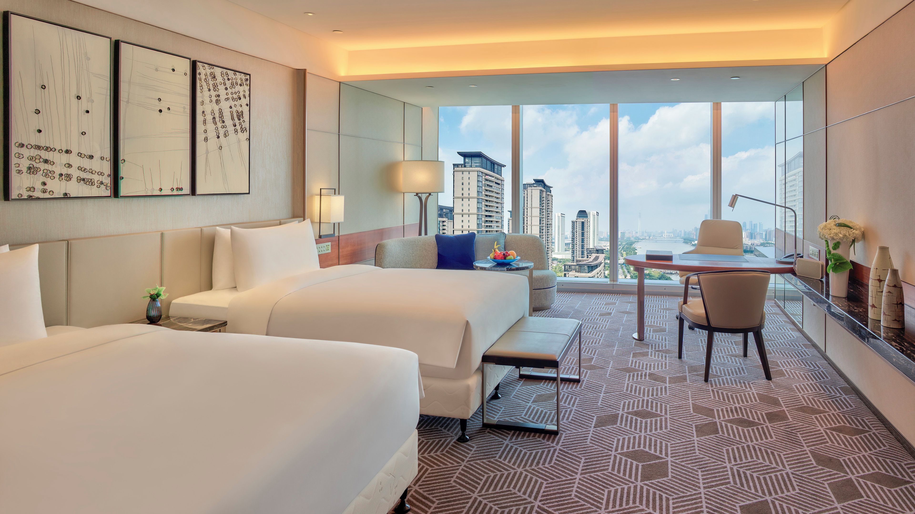 Ein Hotelzimmer mit zwei Betten, einem Schreibtisch, einem Stuhl, einem großen Fenster mit Stadtblick und Bildern an der Wand.