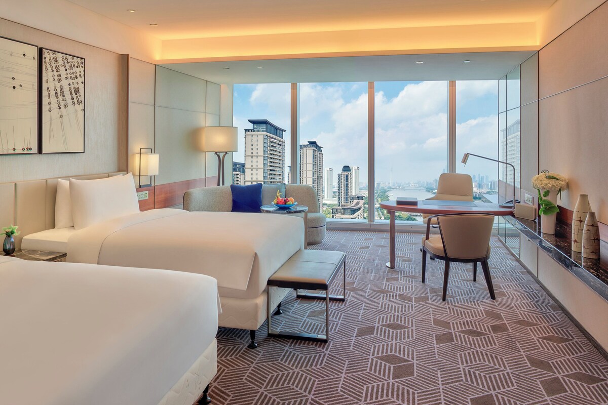 A hotel room with two beds, a desk, a chair, a large window with a city view, and artwork on the wall.