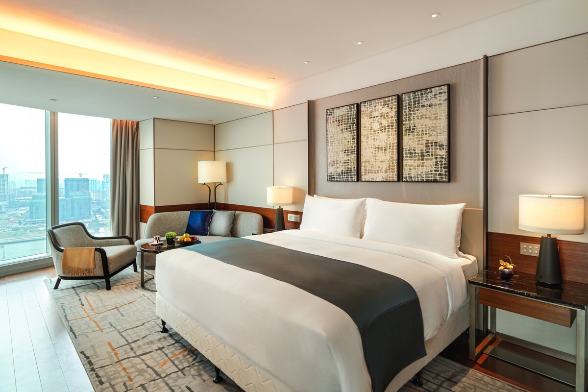 A modern hotel room with a large bed, a desk, a chair, and a view of the cityscape.