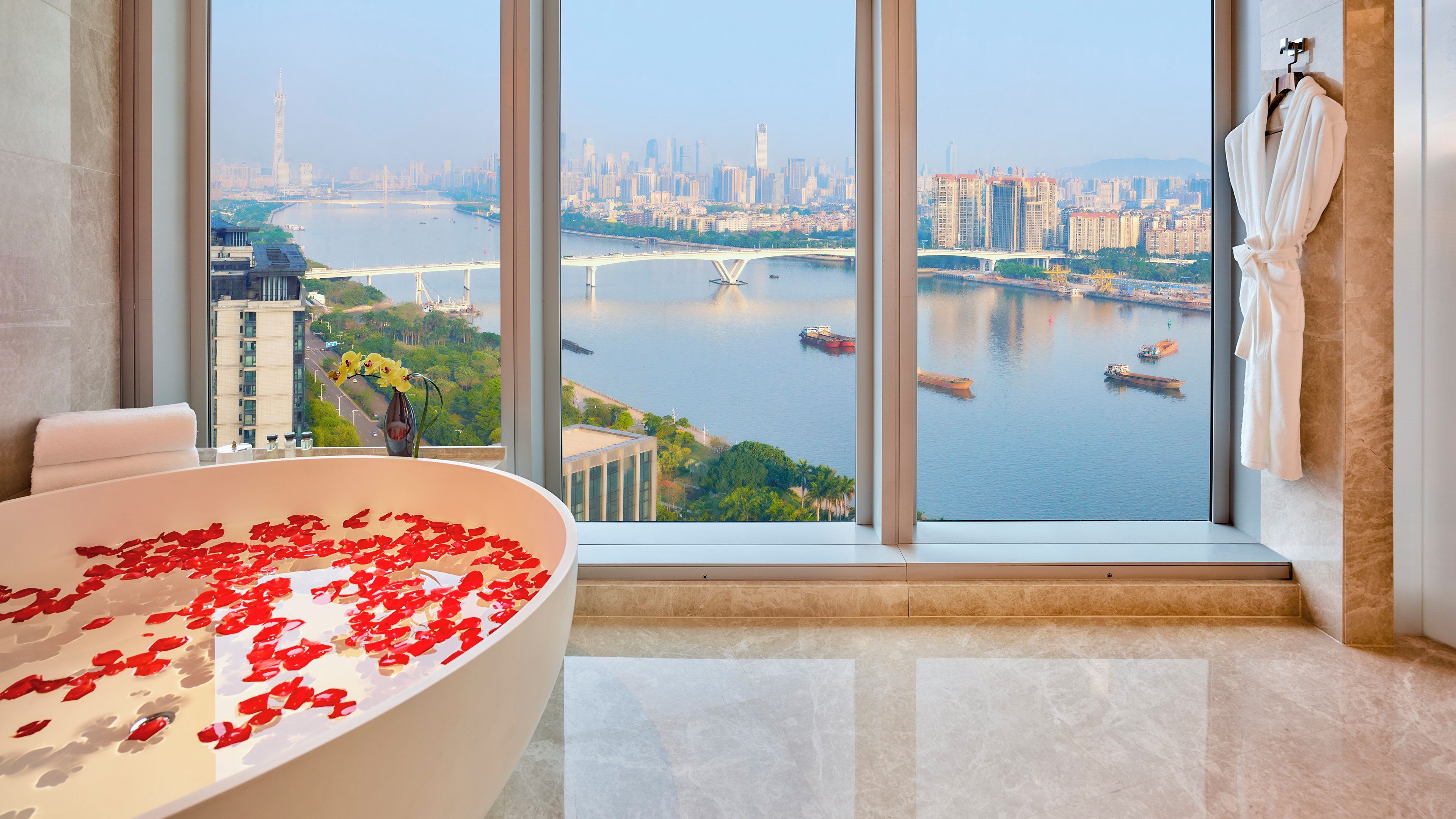 A modern bathroom with a large bathtub filled with rose petals, a view of a cityscape through large windows, and a robe hanging on the wall.