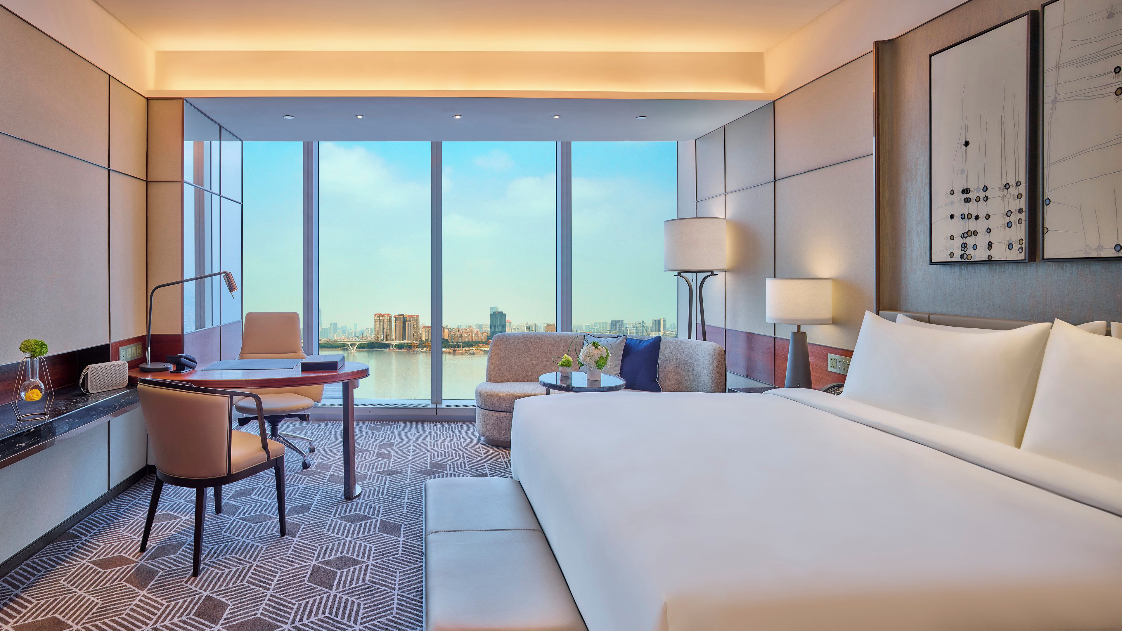 Habitación de hotel con una cama grande, un escritorio con silla, una mesita de noche con lámpara y vistas a la ciudad a través de amplias ventanas.