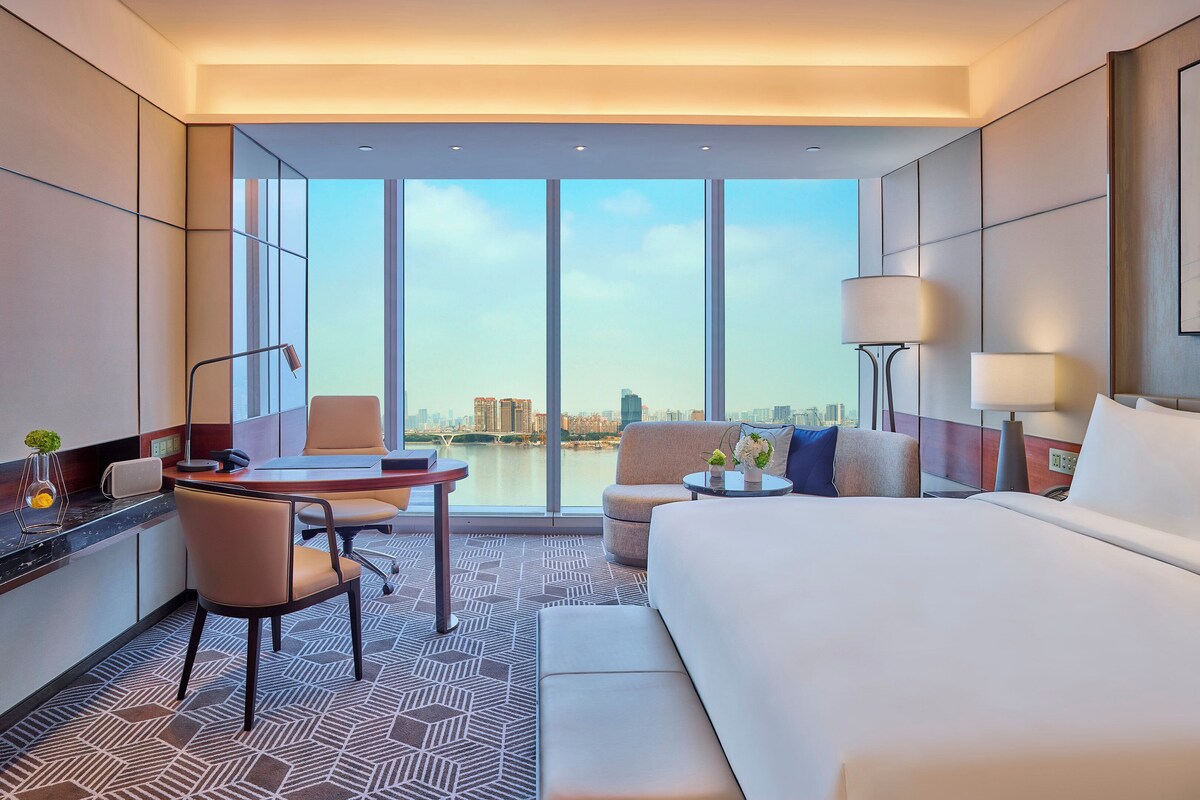 A hotel room with a large bed, a desk with a chair, a small table with a lamp, and a view of a cityscape through large windows.