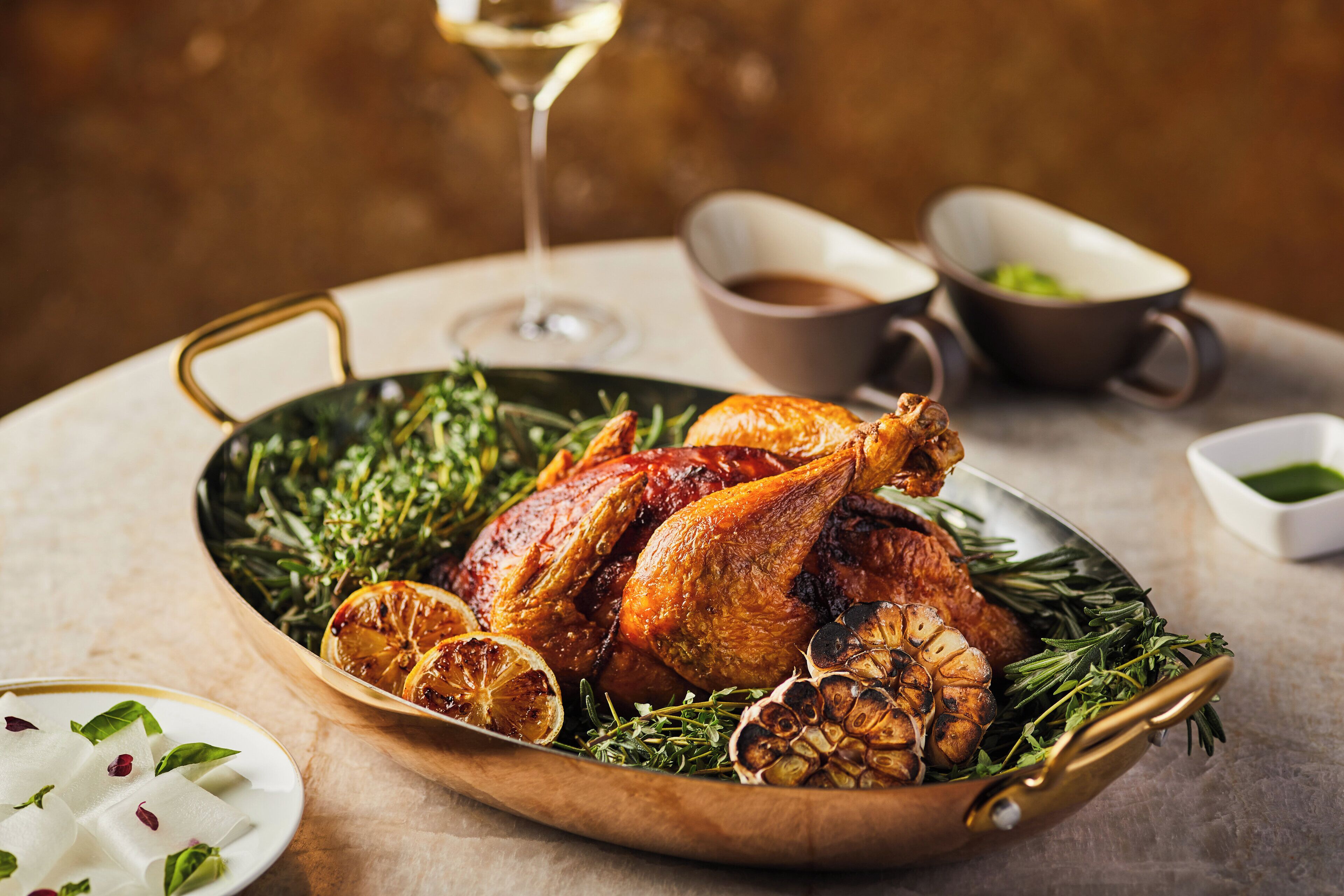 Une dinde rôtie, présentée sur un grand plat, garnie d’herbes aromatiques et de tranches d’agrumes, accompagnée d’une sauce et d’un verre de vin blanc.