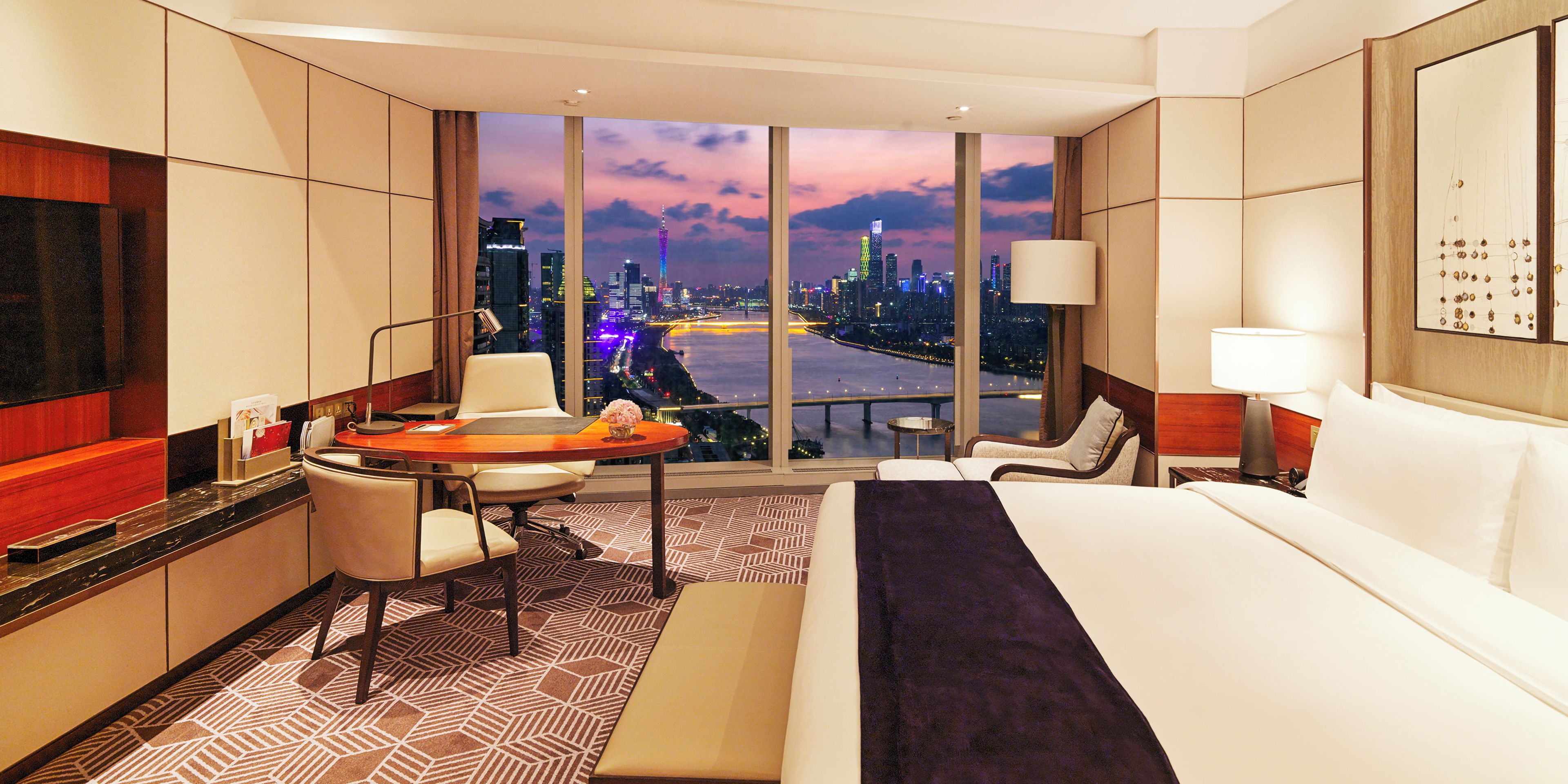 A hotel room with a large bed, a desk, a chair, a small table, and a view of a cityscape at dusk.