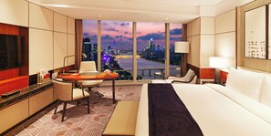 A hotel room with a large bed, a desk, a chair, a small table, and a view of a cityscape at dusk. - InterContinental Guangzhou Exhibition Center by IHG (Guangzhou)