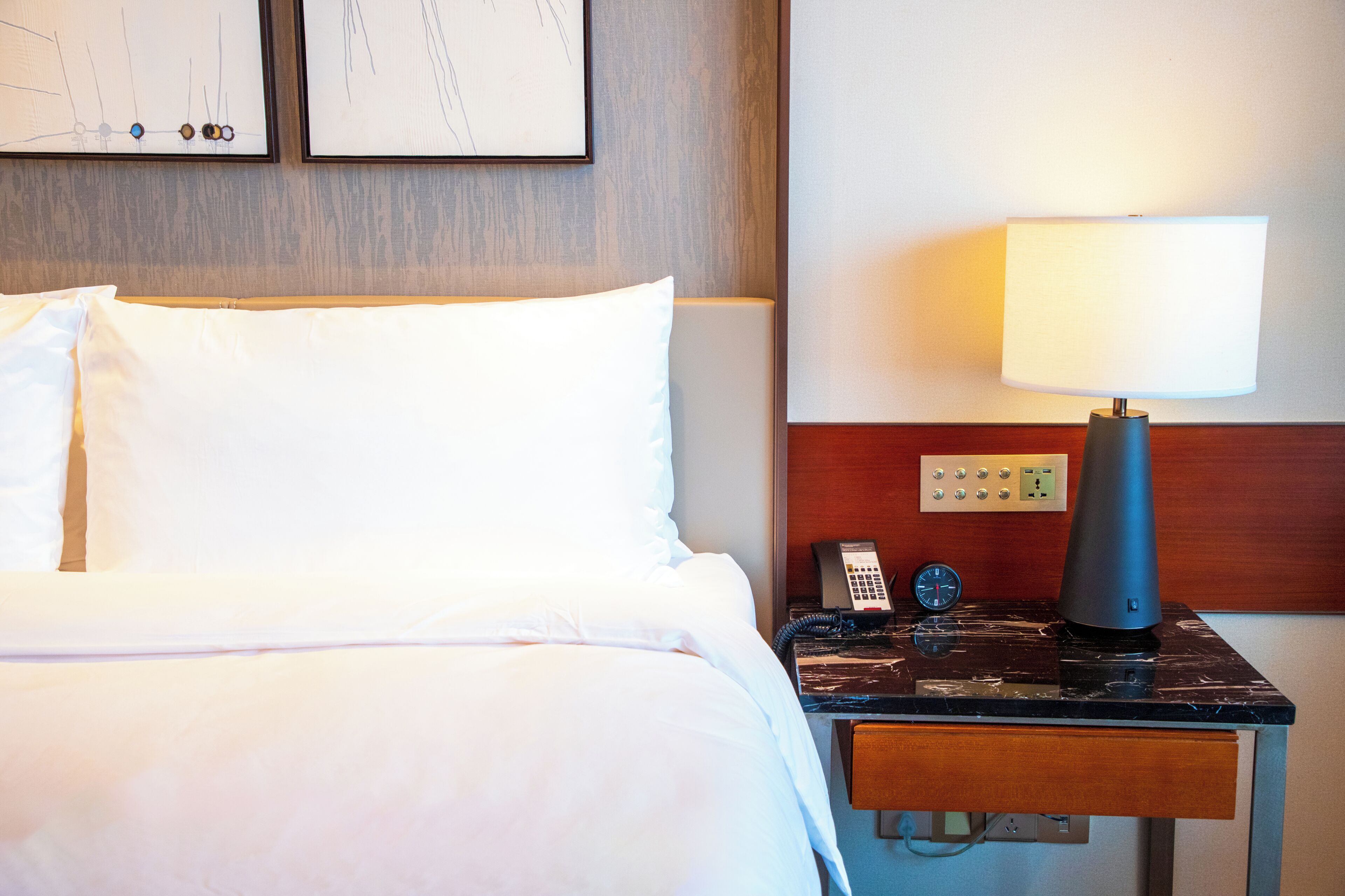 A hotel room with a bed, a nightstand with a lamp, a phone, and a clock.
